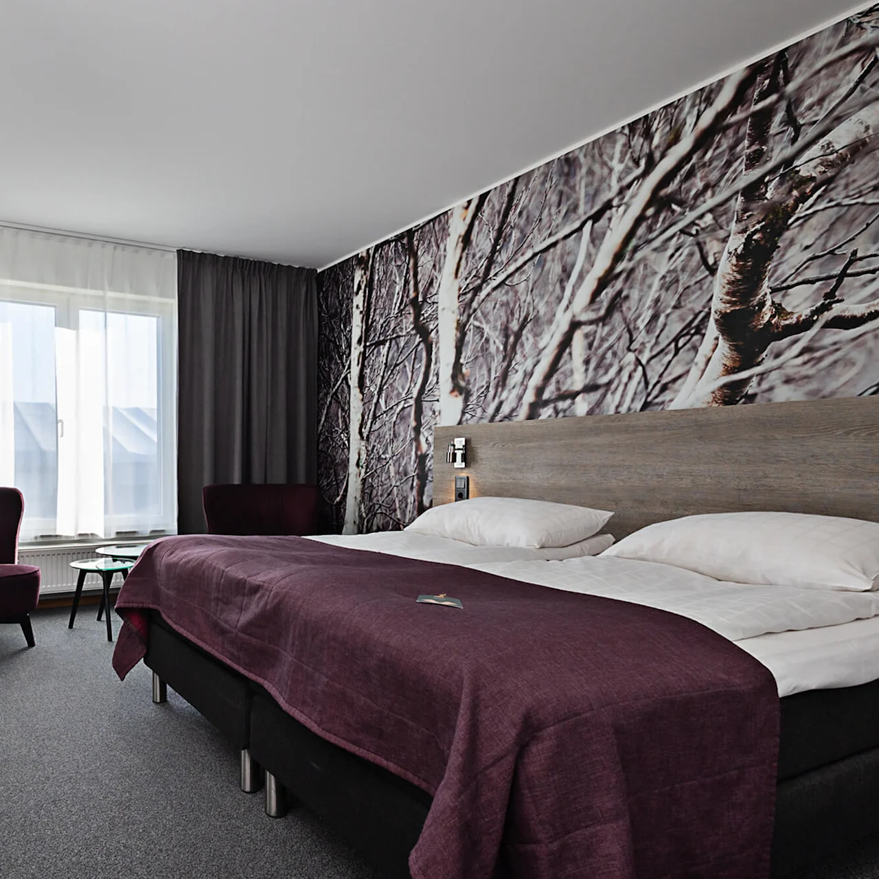 Ett hotellrum med en dubbelsäng täckt med lila sängkläder, flankerad av stolar. Ett fönster med gardiner lyser upp skogstematapeten, vilket skapar en mysig atmosfär.