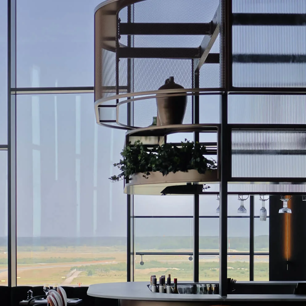 A modern, circular bar with high chairs and bottles sits in an open space with large windows overlooking a scenic landscape, featuring lush green foliage as decor.