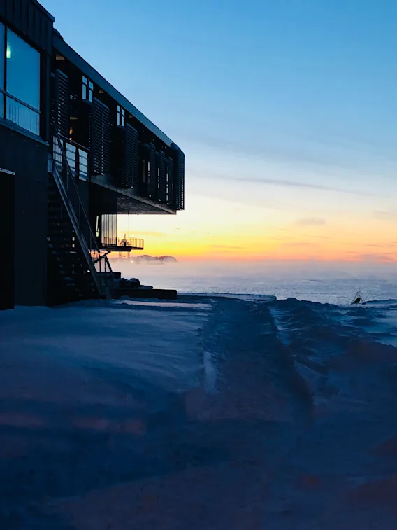En moderne bygning strekker seg over snødekt bakken, med utsikt over et tåkete hav ved solnedgang. Himmelen går fra varm oransje til dyp blå og skaper en rolig, kjølig atmosfære.
