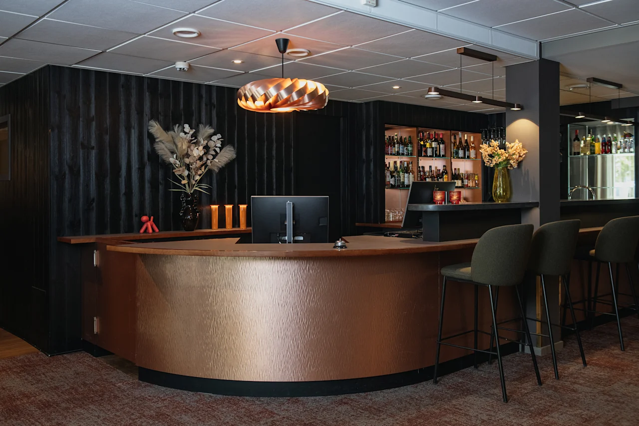 Et moderne hotellresepsjons- og barområde med en buet kobberdisk, barkrakker og hyller fylt med flasker.