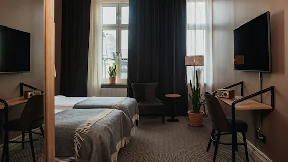 Two neatly made twin beds with gray throws are placed in a hotel room featuring a wall-mounted TV, dark curtains, a small work desk, chair, and large potted plants.