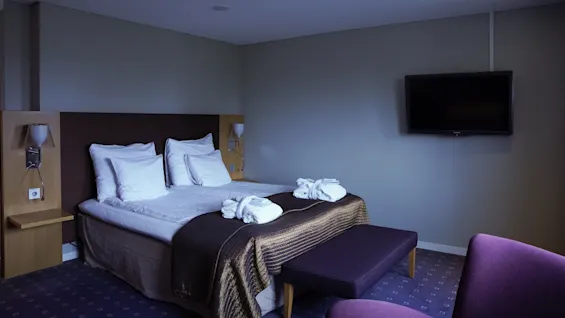 A neatly made double bed in a hotel room with a flat-screen TV on the wall and a sitting area.