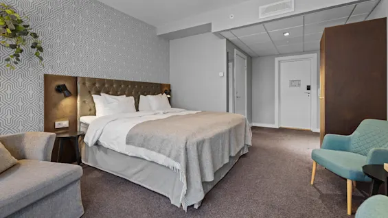 Ett modernt hotellrum med en stor säng, två fåtöljer och en mönstrad accentvägg, som erbjuder en bekväm och inbjudande plats.