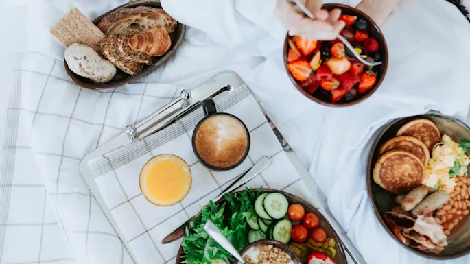 Frokost på sengen med frisk frukt, croissanter og et utvalg av bakverk og brød på en rutet duk, ledsaget av kaffe og appelsinjuice, skaper en koselig og innbydende setting.