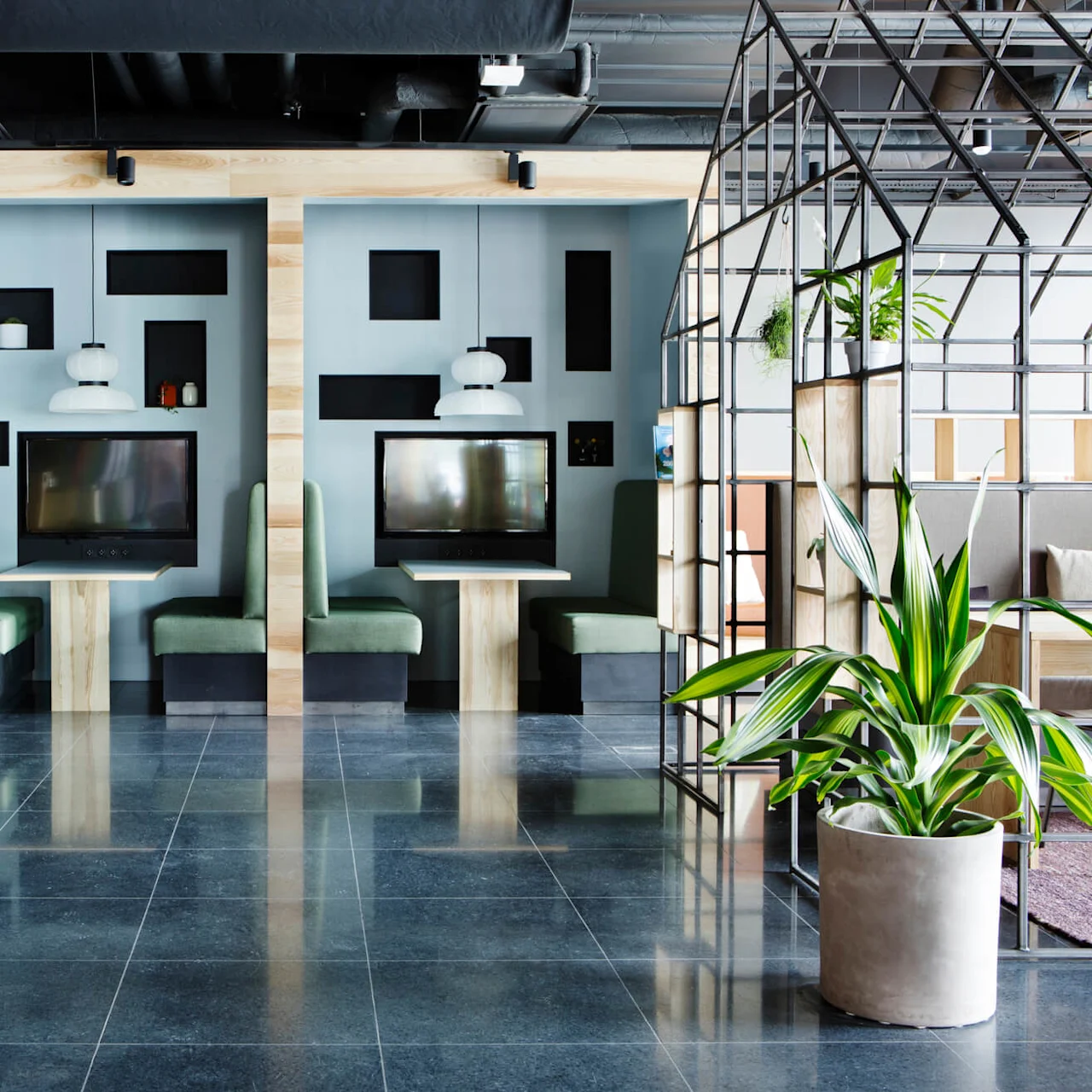 En modern kontorslounge med snygga möbler, tv-apparater på stativ, växtdekorationer och en geometrisk rumsavdelare i ett rymligt, välbelyst inre.