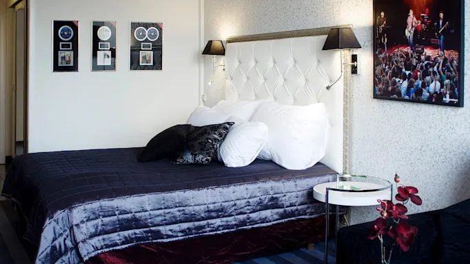 Siististi pedattu sänky koristetyynyineen tuftatun päädyn edessä, ympäröitynä musiikkiteemaisella seinäkoristeilla, viihtyisässä, modernissa huoneessa.