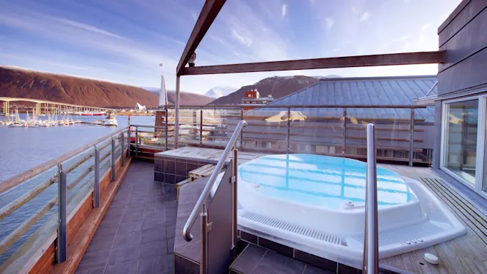 A rooftop hot tub overlooking a marina with boats, set against a backdrop of mountains and blue skies.