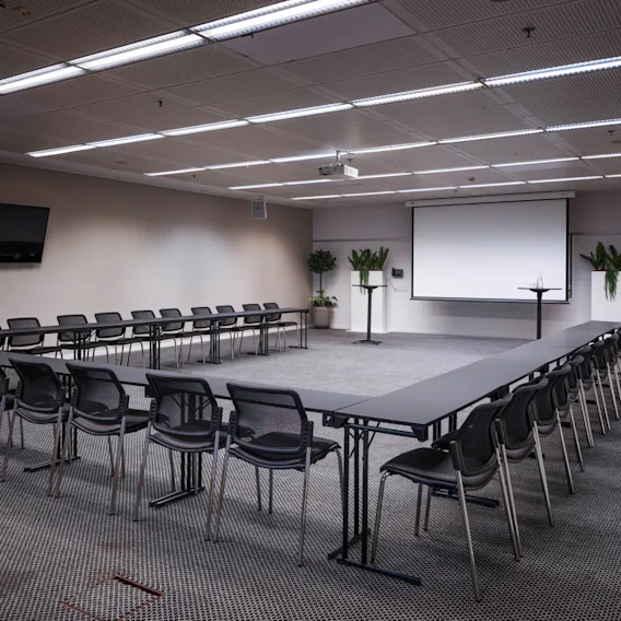 A conference room with U-shaped tables surrounded by black chairs. A projector screen and television are mounted on the wall. Several plants decorate the space.