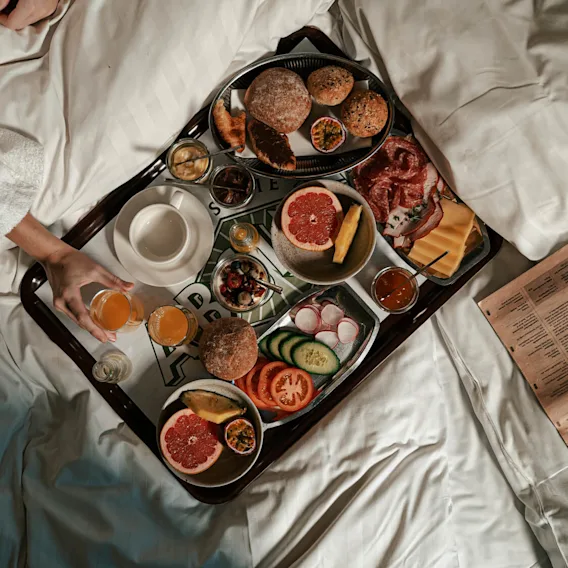 En frukostbricka med bröd, grapefrukt och kött på en säng. En person i morgonrock håller ett glas juice. En tidning ligger bredvid. Text: ”State”.