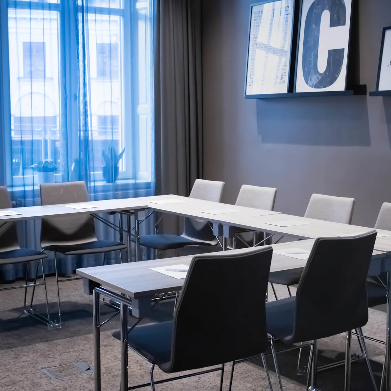 Et moderne konferanserom med langt bord, flere stoler, og store vinduer med tynne gardiner; sollys filtreres gjennom og fremhever rommets enkelhet og profesjonalitet.