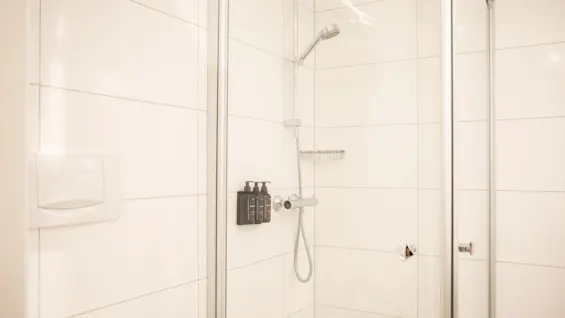 Shower enclosed by glass panels, featuring mounted dispensers and a handheld showerhead. White tiled walls provide a clean, modern bathroom setting.