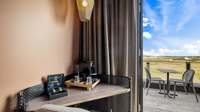 Espressomaskin og kopper på et lite trebord ved siden av en stol; plassert nær et vindu med beige gardiner. Utenfor er det en balkong med et bord som vender mot et bredt landskap under en klarblå himmel.