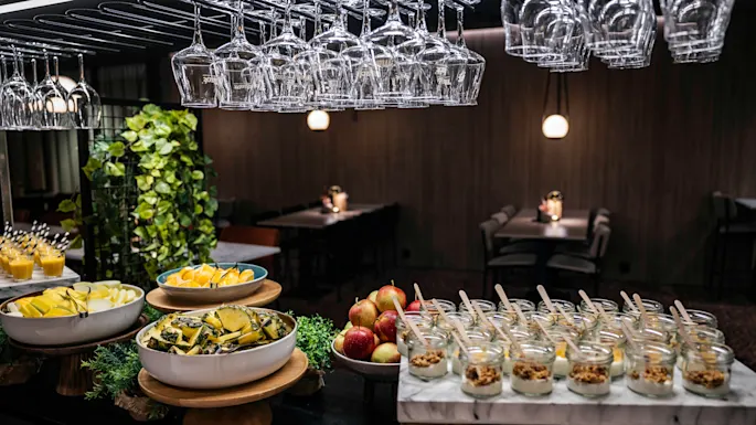 Glasses hang above a buffet table with fruit bowls and yogurt jars, surrounded by indoor plants. Dimly lit dining tables are visible in the background.