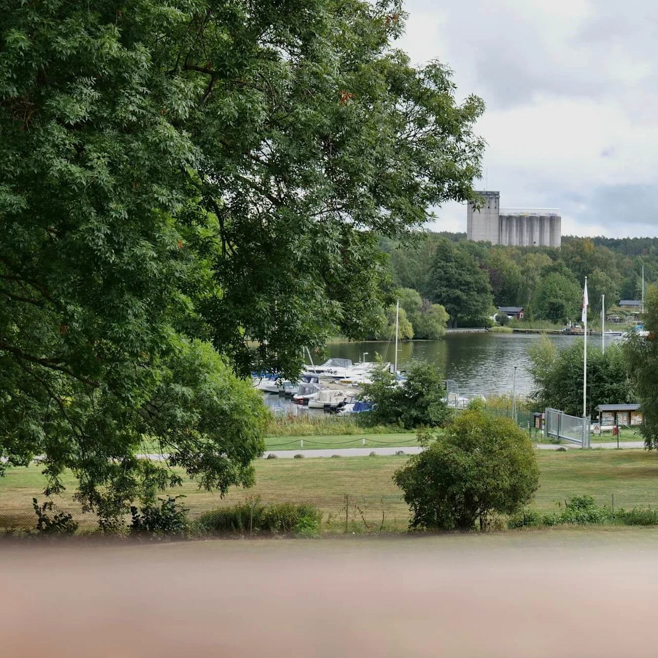 Både til kaj ved en marina, hvilende på stille vand, omgivet af frodige grønne træer under en skyet himmel, med industrielle siloer synlige i det fjerne.