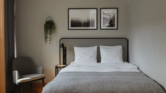 En pent oppredd seng med hvite puter og et grått teppe står i et lite rom. Innrammede bilder henger over, og en hengende plante er ved veggen ved siden av en koselig stol.