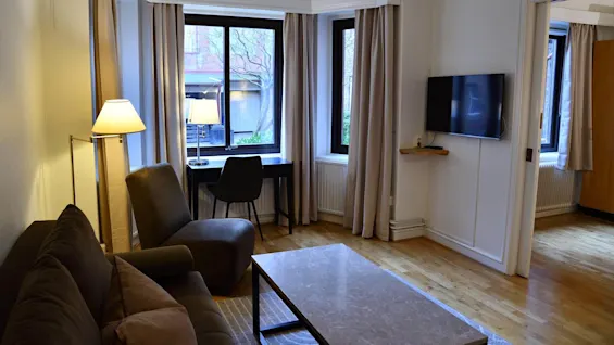 Olohuone, jossa sohva ja tuoli sohvapöydän edessä. Vieressä on lamppu. Ikkunan ääressä on työpöytä ja tuoli. Televisio on seinällä.