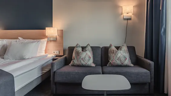 A bed and sofa sit in a dimly lit hotel room, illuminated by wall lamps. The bed has white pillows, and the gray sofa features patterned cushions. A small table is in front.