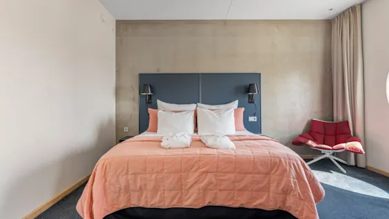 Et hyggeligt hotelværelse med en stor seng, ferskenfarvet sengetæppe, hvide puder, to badekåber og en moderne rød lænestol.