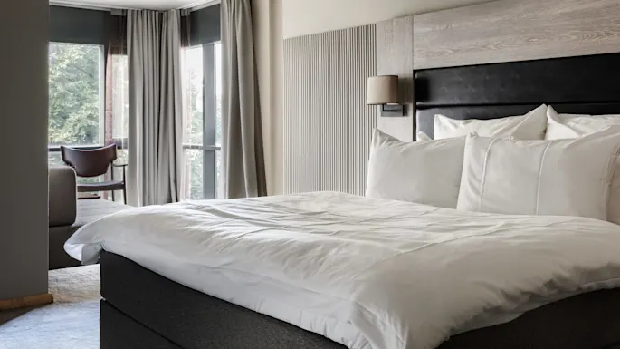 A neatly made bed with white linens and pillows, positioned in a modern hotel room with large windows, draped gray curtains, and a chair in the corner near a sofa.