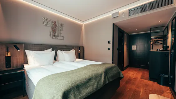 A modern hotel room with a comfortable bed, bedside table, and an open wardrobe area, featuring warm lighting and wooden floors.