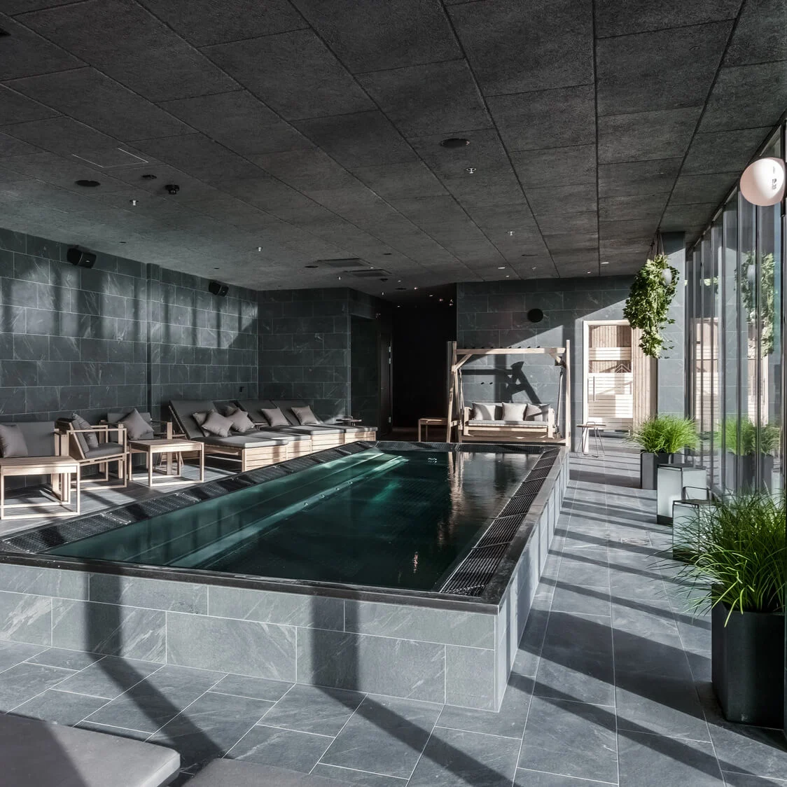 En rektangulær indendørs swimmingpool med klart vand, omgivet af polstrede træmøbler, i et moderne stenrum med store vinduer og grønne planter.