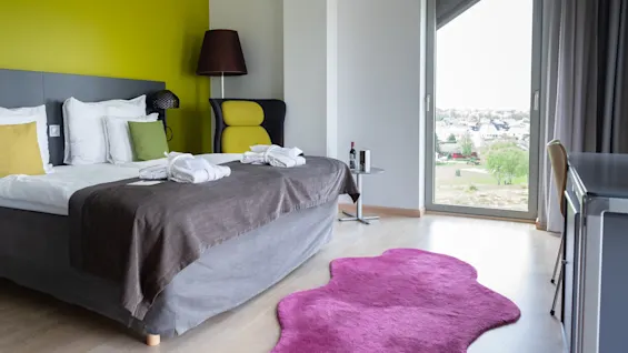 Ett modernt hotellrum har en dubbelsäng med vita lakan, en grön accentvägg och ett fönster med utsikt över byggnader. En rosa matta och en fåtölj kompletterar inredningen.