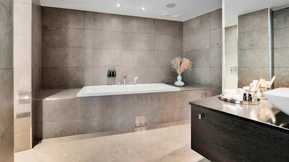 A bathtub is the focal point in a modern bathroom, surrounded by gray tiles. A countertop with a white sink, a tray of toiletries, and decorative vases provide context.