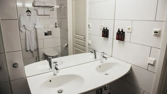 Et hvidt badeværelse har en dobbeltvask med et spejl, der reflekterer et toilet og hængende badekåber. Flisevægge har to monterede sæbedispensere. Håndklæder hviler på en hylde over.