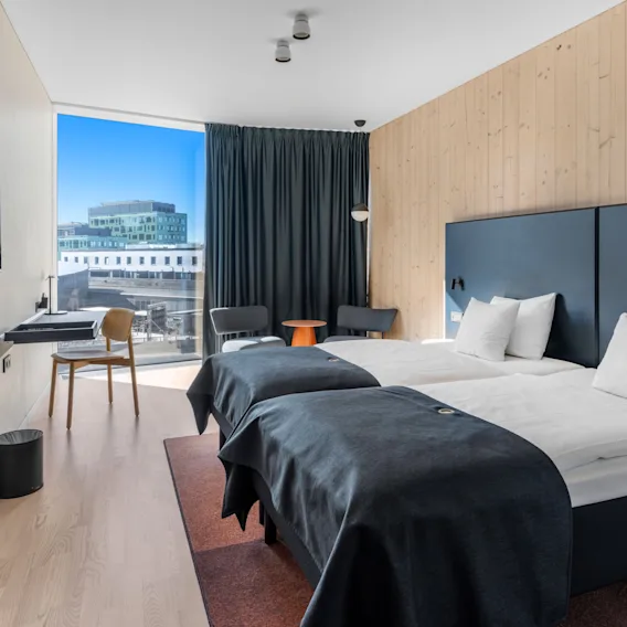 Ljust och modernt hotellrum med två sängar, trägolv och ett stort fönster med utsikt över staden.