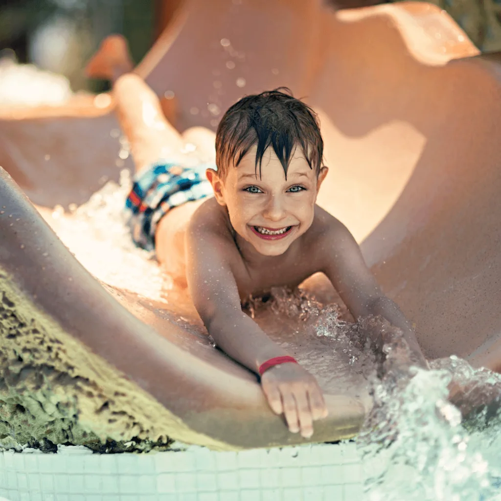 En ung gutt sklir nedover en våt vannsklie, smilende med spenning. Han har på seg fargerike badebukser og er omgitt av sprutende vann i et solfylt utendørs badeland.