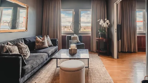 A living room with a dark sofa adorned with patterned pillows faces a marble coffee table holding a candle. Large windows with curtains showcase a bright sky view.