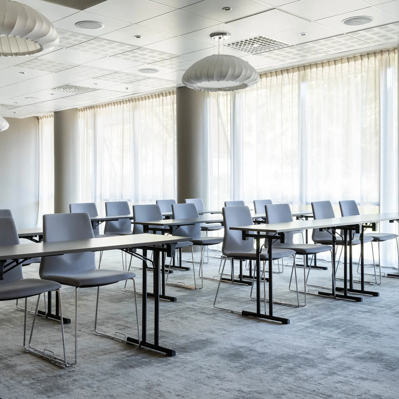 Classroom with rows of gray chairs and desks facing windows. Ceiling lights hang overhead, and soft natural light filters through sheer curtains. The view is from an open doorway.