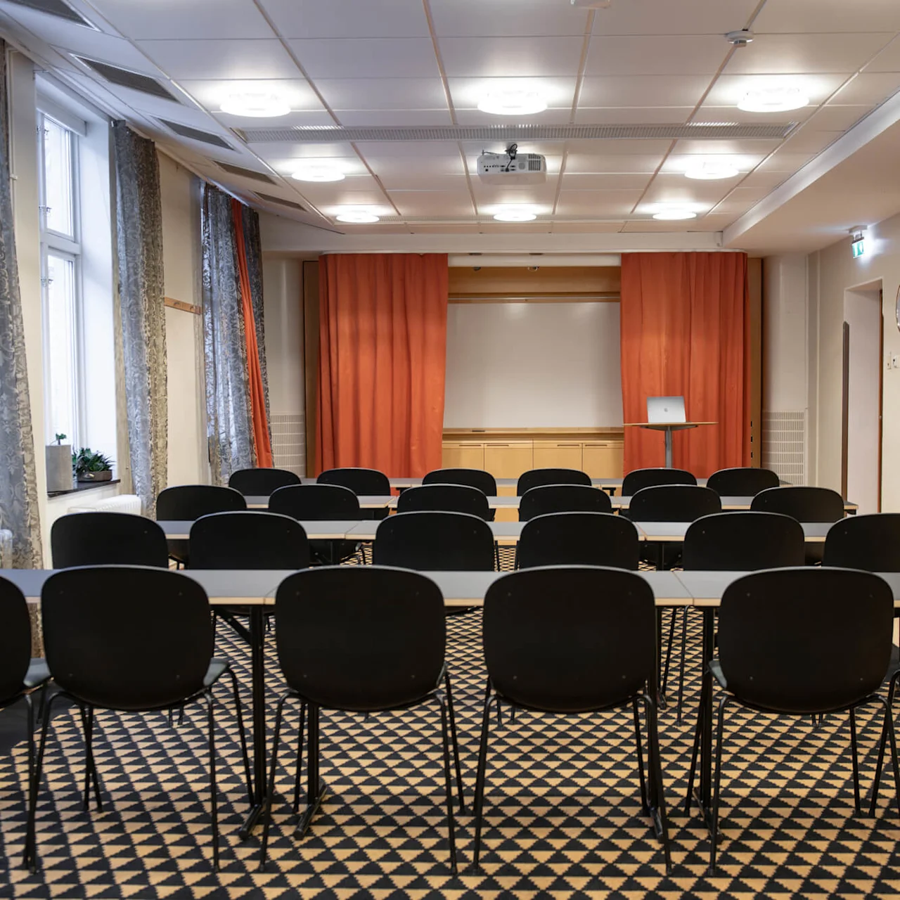 Rows of black chairs face a whiteboard flanked by red curtains. A laptop sits on a stand, with windows and patterned carpet creating a formal presentation setting. 