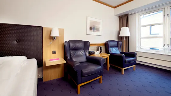 Et pænt arrangeret hotelværelse med en seng og to lænestole oplyst af væglamper, og et indrammet billede over stolene.