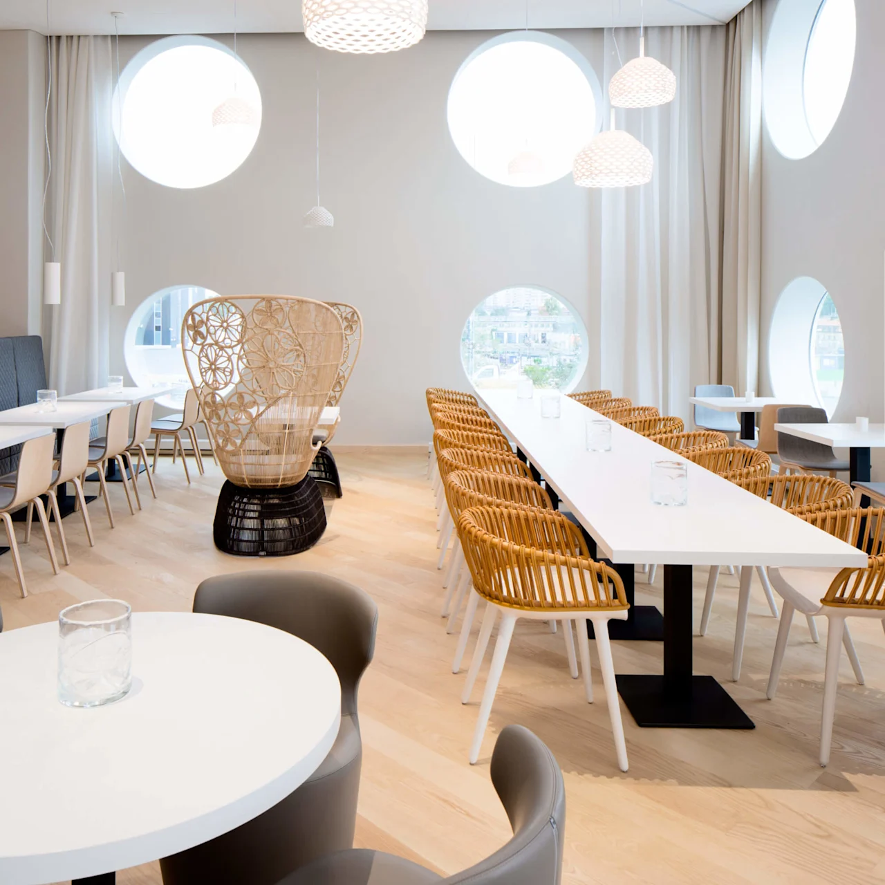 Hvide borde og stole er arrangeret i en moderne spisestue med cirkulære vinduer og pendellamper. En flettet stol står mod bagsiden, i et minimalistisk miljø.