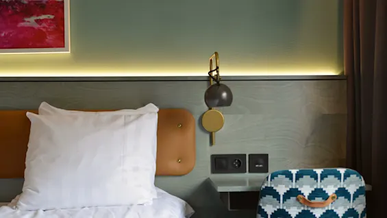 Seng med hvid pude ved siden af en mønstret stol, under en moderne væglampe i et stilfuldt hotelværelse. Et abstrakt maleri dekorerer væggen ovenfor.