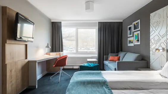 Ett hotellrum med en säng täckt av turkost sängkläder, en väggmonterad TV, en orange stol vid ett skrivbord och en grå soffa vid ett fönster med utsikt över parkerade bilar.