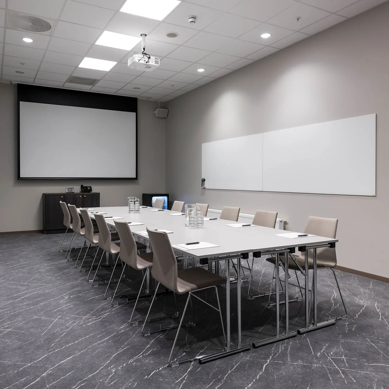 Konferansebord med åtte stoler, notisblokker og vannmugger i et moderne møterom. En tom skjerm og whiteboard er montert på vegger, med projektor over.