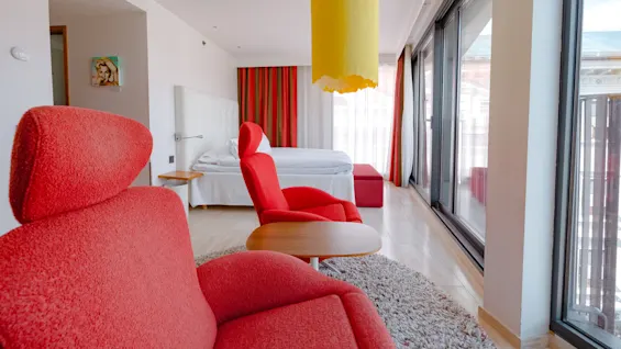 Red armchairs face a bed with a white headboard, beneath a yellow hanging lamp. Large windows line one side, and red-striped curtains add color. A small table sits nearby.