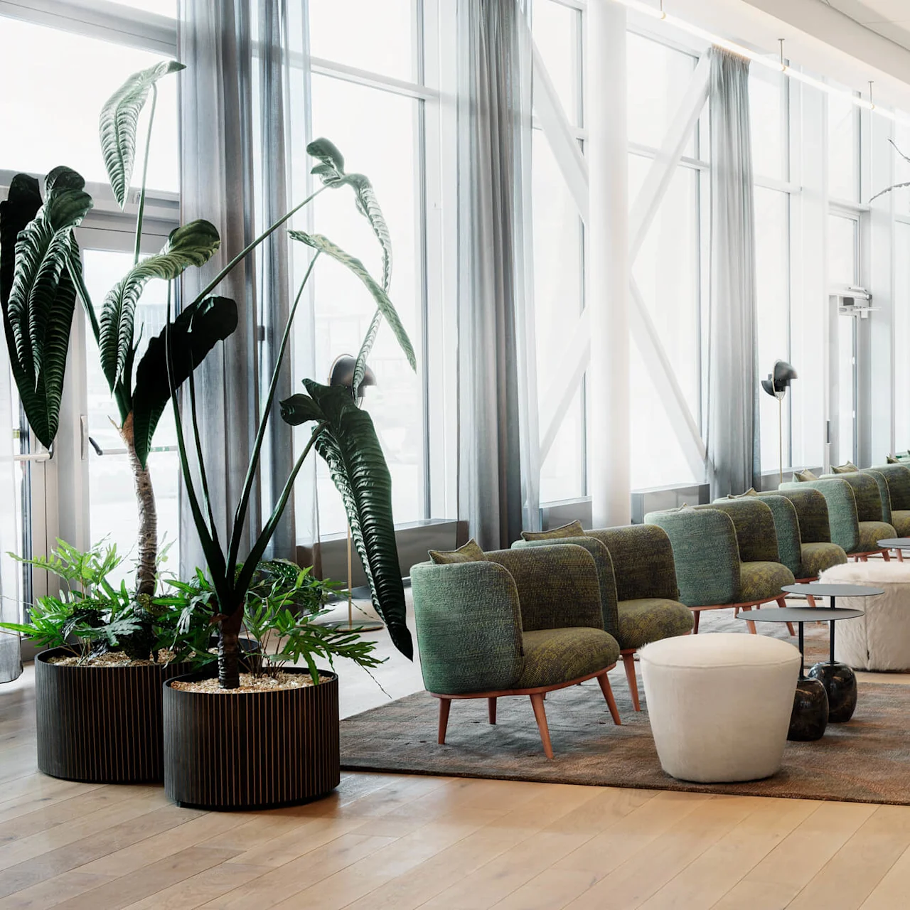 Grønne stoler står på rekke i en lys og romslig lounge med store vinduer, planter og moderne belysning, som skaper en innbydende atmosfære.