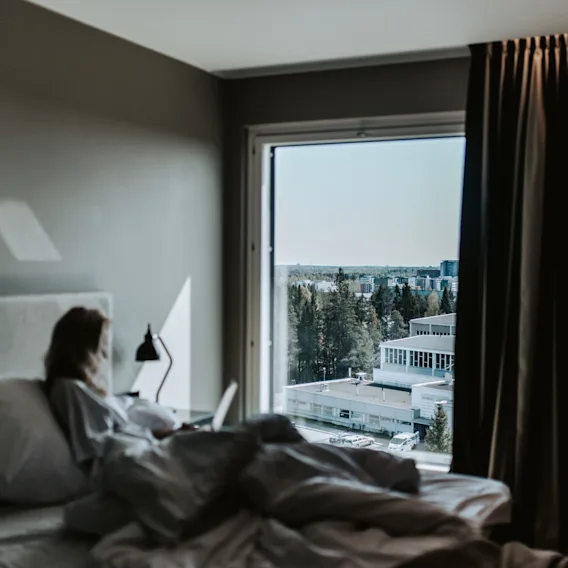 Joku sängyssä kannettavan tietokoneen kanssa, katselee ikkunasta kaukaista kaupunkia ja puita aurinkoisena päivänä.