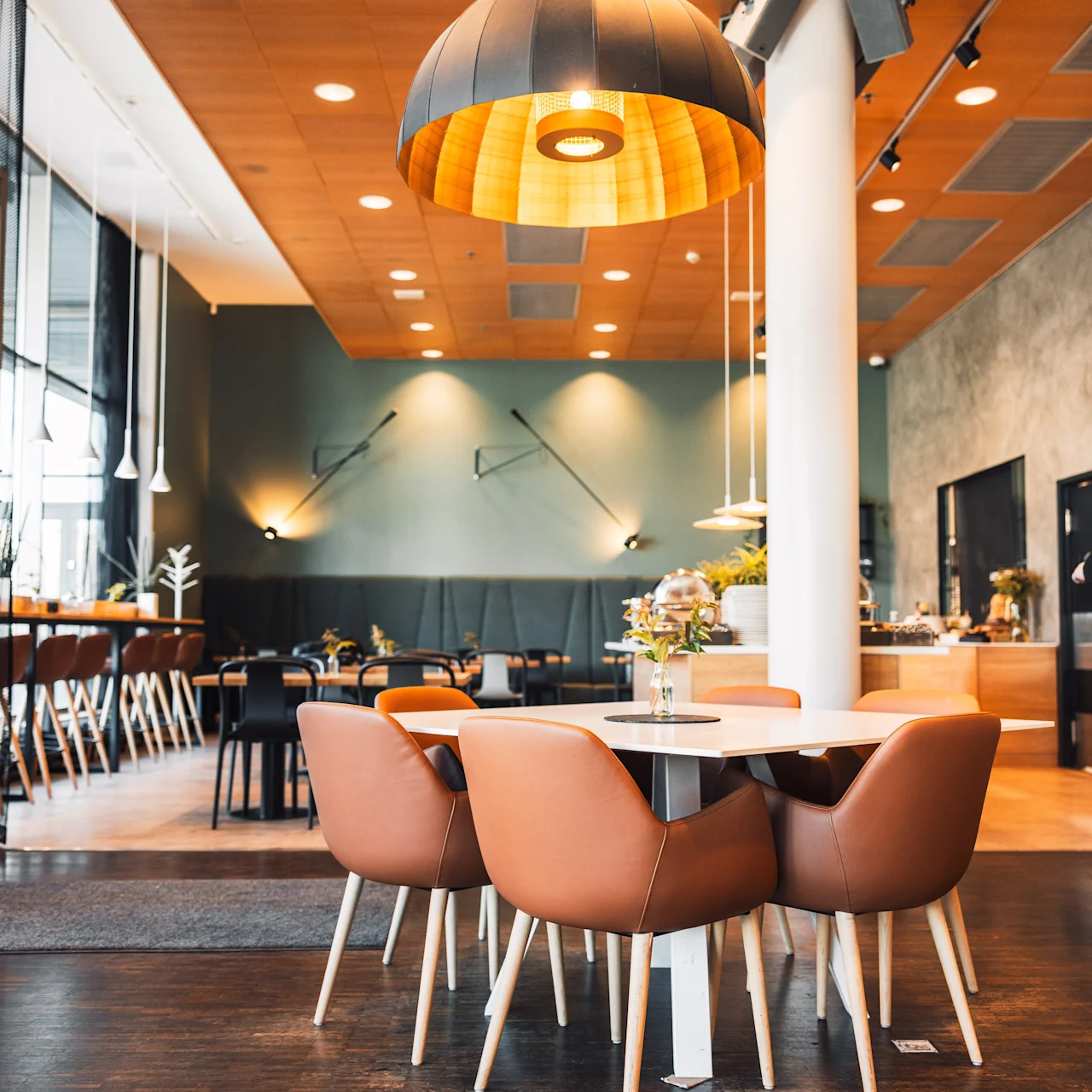 A round table with four brown chairs stands under a large hanging light in a modern, stylish cafe with wooden floors and minimalistic decor.