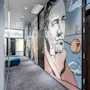 A hallway with a mural of a man smoking a pipe, surrounded by modern design elements. Text includes "ROCKIN'" on a door and "SATISFACTION" reflected on glass.
