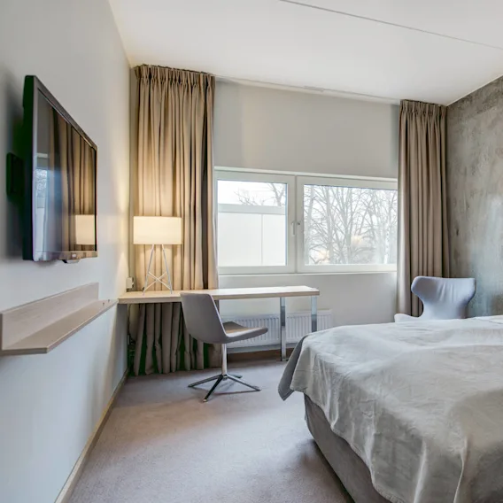 Et pent innredet hotellrom med dobbeltseng og grått sengeteppe. Rommet har en veggmontert TV, skrivebord med lampe, stol og et vindu som viser trær utenfor.