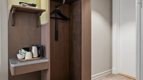 A cozy hotel room corner with a wooden wardrobe, a shelf for personal items, and a convenient coffee and tea station.