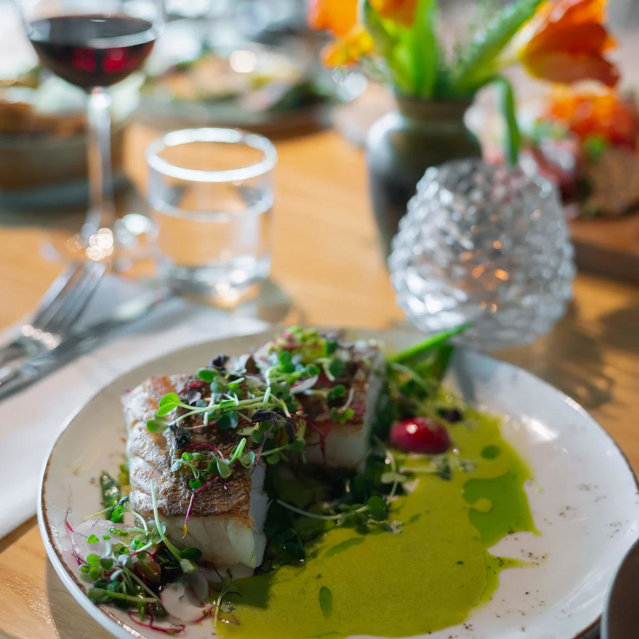 Grillad fisk toppad med mikrogrönt ligger på en vit tallrik med grön sås, tillsammans med ett glas rött vin och vatten på ett träbord med blommor.