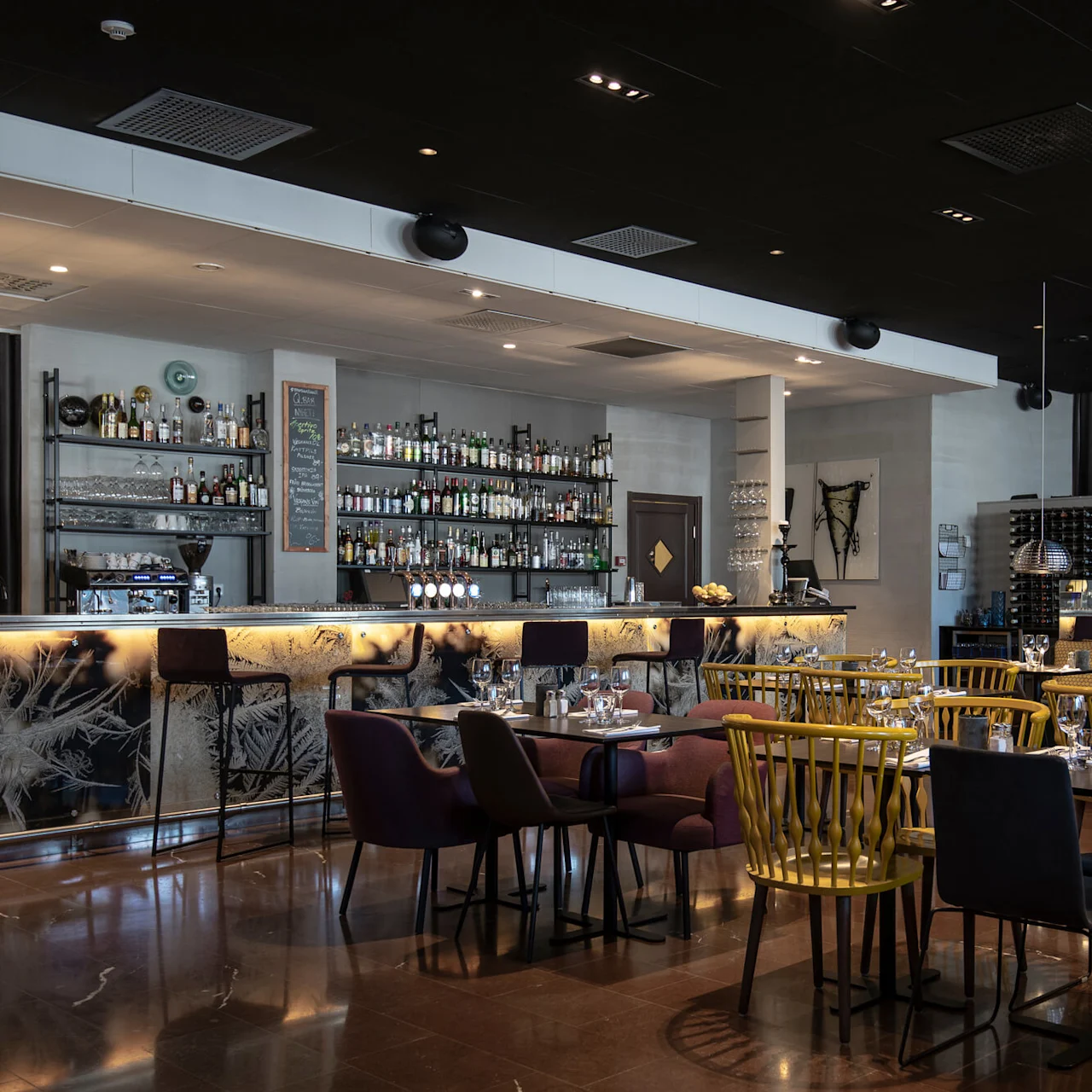 En rymlig bar med upplysta diskar och hyllor fyllda med flaskor; olika bord och stolar fyller den moderna, varmt upplysta restaurangmiljön.