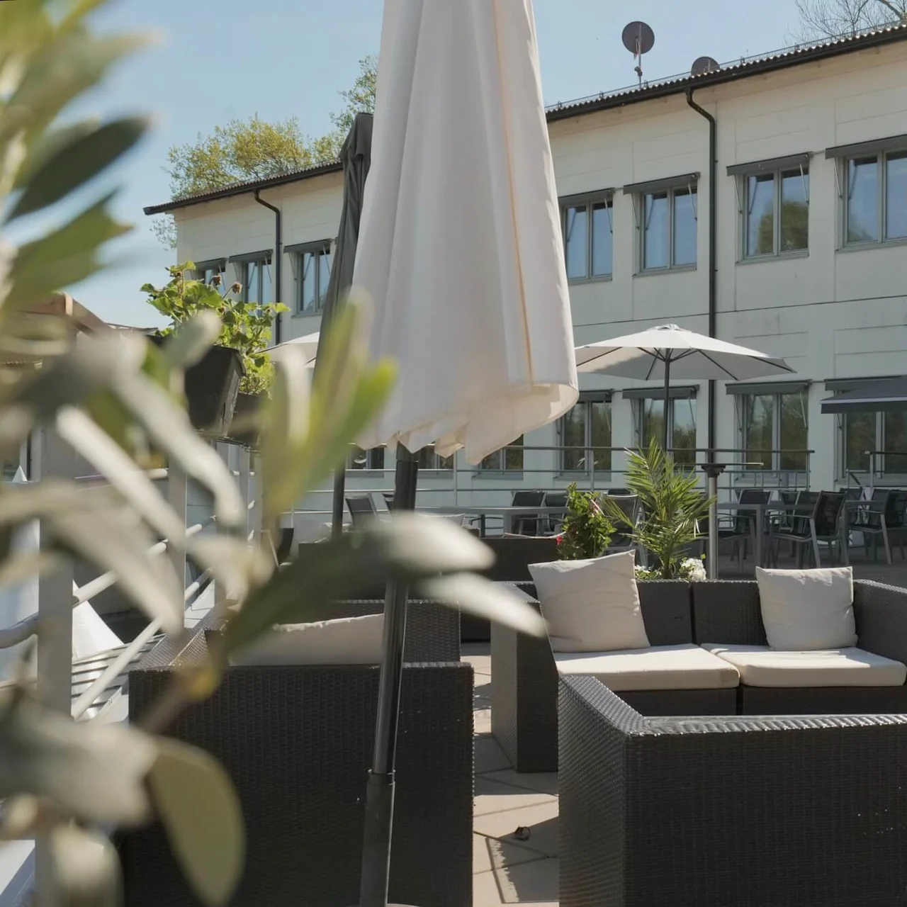 Udendørs møbler står på en terrasse omgivet af potteplanter og store paraplyer. Scenen er sat mod en beige bygning med mange vinduer.