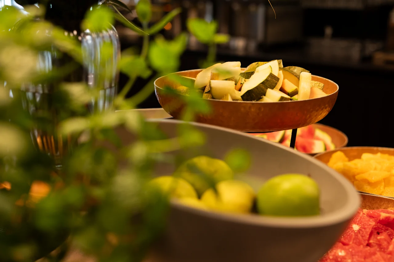 Et levende nærbilde av en fruktbuffé med skåler med fersk melon, vannmelon og andre frukter, innrammet av grønne planter.