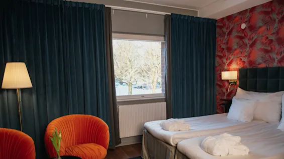 A hotel room with twin beds neatly arranged under a vibrant red-patterned wall. Cozy orange chairs and a lamp enhance the space beside a window showing snowy trees.